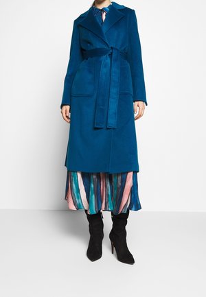 Femme portant un long manteau bleu sarcelle ceinturé par-dessus une robe à rayures multicolores et des bottes à talons noires, debout devant un fond blanc uni.