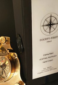 Handcrème tube in een zwarte doos met witte etikettering; heeft een gouden detail dat lijkt op een kompas aan de zijkant. Tekst luidt "Crema Mani EXTRA REPAIR."