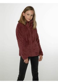 Giacca in pile bordeaux con colletto alto e zip frontale, caratterizzata da una finitura morbida e testurizzata e maniche lunghe. Abbinata a jeans neri.