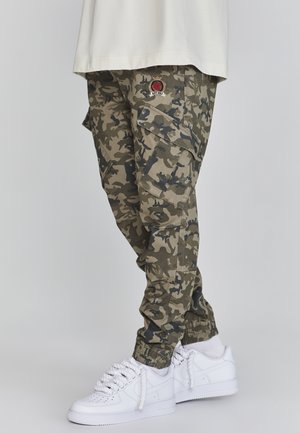 Benedenlichaam gekleed in groene camouflage cargobroek met elastische boorden en witte sneakers met dikke veters, tegen een effen achtergrond.