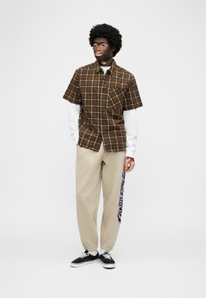Νεαρός με σκούρο δέρμα και σγουρά μαλλιά, φοράει καρό καφέ κοντομάνικο πουκάμισο πάνω από λευκή μακριά μπλούζα, μπεζ παντελόνι με σχέδιο στο πλάι και μαύρα αθλητικά παπούτσια.