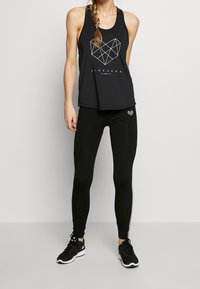 Svart linne med geometrisk hjärtformad design och vit text, tillsammans med svarta leggings med en liten hjärtlogotyp. Svarta träningsskor.