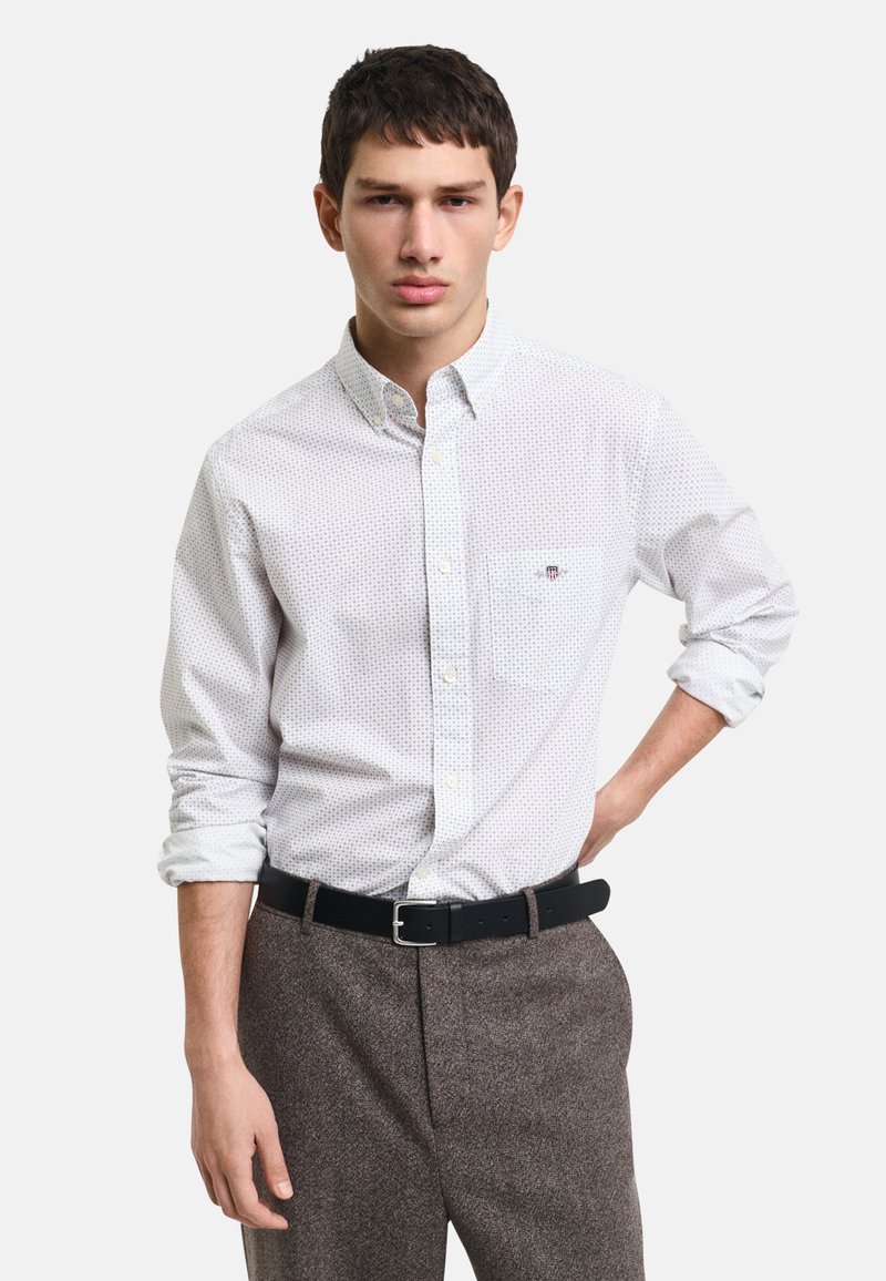 Camisa blanca de botones con un pequeño estampado, con un bolsillo en el pecho y un cuello. Combinada con pantalones marrones texturizados y un cinturón negro.