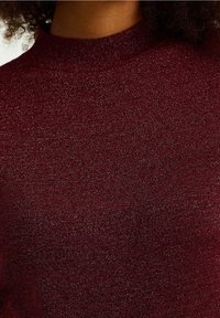 Burgunderfärgad stickad tröja med hög halsringning, som har en subtil metallisk glans och en slät yta. Närbild av tyget.