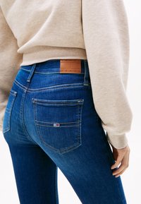 Tamsiai mėlyni džinsai iš džinso su lygiu paviršiumi; turi odinę etiketę ant juosmens nugaros pusėje ir galines kišenes su siuvimo detalėmis.