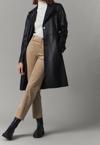 Svart läder trench coat över en vit turtleneck, kombinerad med beige kordfluwelpants och svarta ankelstövlar. Slät textur och rena linjer.