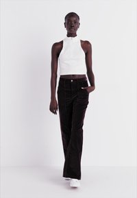Witte mouwloze top met hoge kraag, gecombineerd met donkerbruine corduroyen broek. Model staat met één hand in de zak en draagt witte sneakers.