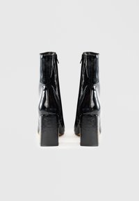 Botines cortos de charol negro con un acabado liso, tacones cuadrados altos y cierre de cremallera en la parte trasera.