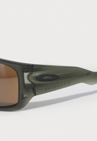 Ochelari de soare verzi transparenți cu lentile brunii, având un design curbat și un logo negru proeminent pe bratul ochelarilor. Textură netedă.