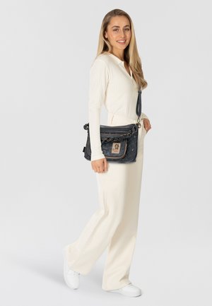 Marineblaues, gesteppte Umhängetasche mit beige Farbakzenten, silberner Kettenumhängeleiste und einem Logo-Patch. Das Modell trägt ein weißes Oberteil und cremefarbene Hose und hält die Tasche.