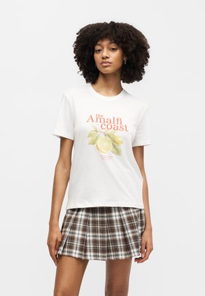 Jonge vrouw met krullend haar die een wit T-shirt draagt met "de Amalfikust" en een citroengrafiek, gecombineerd met een bruine geruite geplooide rok.