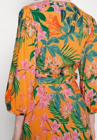 Sukienka w kwiaty z intensywnie pomarańczowego materiału z różowymi i zielonymi tropikalnymi wzorami, z bufiastymi rękawami i wysmuklonym pasem.