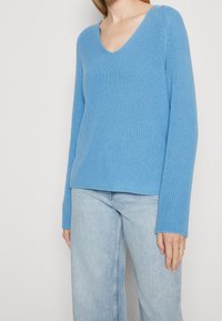 Donna che indossa un maglione a V in maglia blu chiaro e jeans in denim chiaro, in piedi contro uno sfondo semplice.