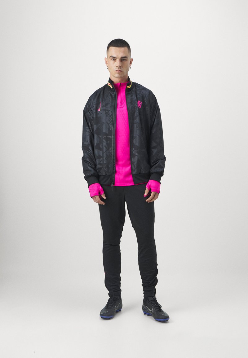 Giacca nera a fantasia con accenti gialli e rossi sopra una maglietta a coste rosa brillante. I pantaloni neri e le scarpe sportive scure completano l'outfit.