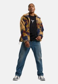Fleece zip-jakke med farverigt blomsterdesign, tan base og marineblå manchetter, parret med sort t-shirt, blå jeans og mønstrede sneakers.