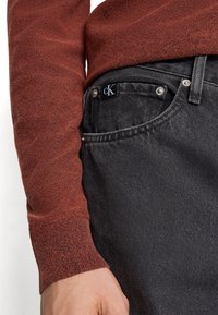 Tmavě šedé džíny z denimu s texturovaným povrchem a stříbrnými detaily, v kombinaci s vínově červeným pleteným svetrem s vroubkovanými manžetami.