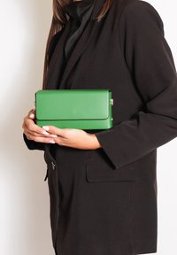 Clutch in pelle verde con forma rettangolare piatta, dotata di un pattino pieghevole e dettagli in metallo dorato, tenuta contro un abbigliamento scuro.