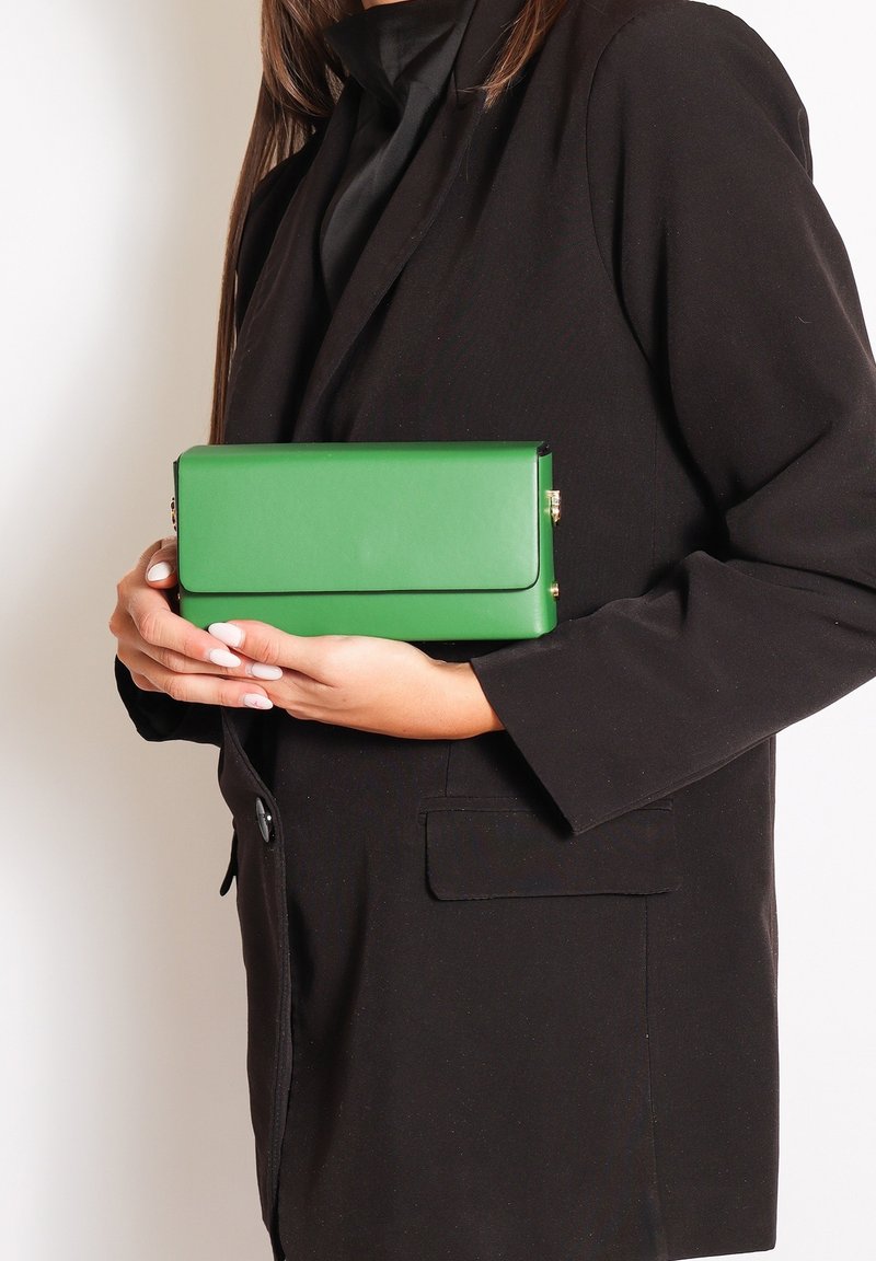 Clutch in pelle verde con forma rettangolare piatta, dotata di un pattino pieghevole e dettagli in metallo dorato, tenuta contro un abbigliamento scuro.