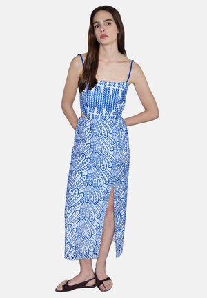 Frau mit langen braunen Haaren, die ein blau-weiß gemustertes ärmelloses Kleid mit Frontschlitz und dunklen flachen Sandalen trägt, steht vor einem schlichten Hintergrund.