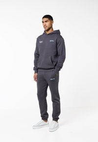 Uomo in piedi che indossa una felpa con cappuccio grigio scuro e pantaloni della tuta abbinati con scarpe da ginnastica bianche su uno sfondo bianco uniforme.