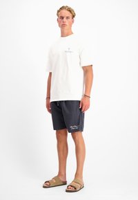 Witte katoenen T-shirt, relaxte pasvorm; donkerblauwe gestreepte shorts met borduursel; bruine sandalen met twee banden. Effen achtergrond.