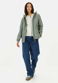 Grüne Steppjacke mit Kapuze, ausgestattet mit einem durchgehenden Reißverschluss und Fronttaschen, getragen über einem hellen Oberteil und lockeren blauen Jeans. Strukturierter Stoff.