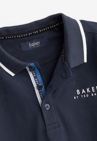 Polo blu navy con rifiniture bianche sul colletto, patta con bottoni a righe blu e bianche e logo "Baker by Ted Baker" sul petto e sui bottoni.