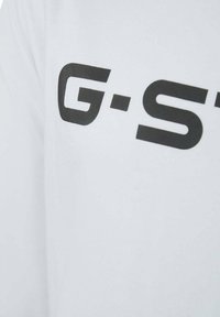 Witte blouse met lange mouwen, voorzien van grote zwarte "G-S" tekst. De stof lijkt soepel en lichtgewicht met een ontspannen pasvorm.