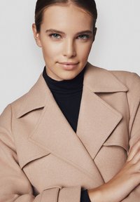 Beżowy płaszcz z wełny z dużymi klapami, o długości pośredniej i subtelnej teksturze. Noszony na czarnym golfie, z luźnymi rękawami.