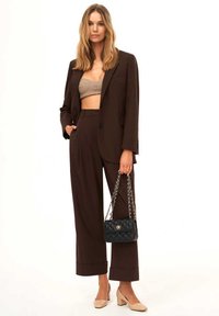 Completo marrone su misura con pantaloni a gamba larga, top corto beige e borsa a tracolla nera trapuntata. Tacchi chunky neutri completano il look.