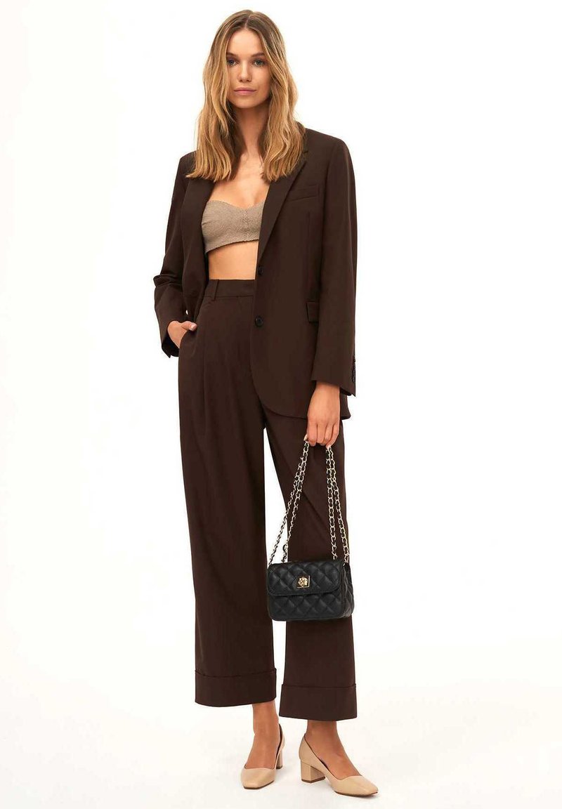 Completo marrone su misura con pantaloni a gamba larga, top corto beige e borsa a tracolla nera trapuntata. Tacchi chunky neutri completano il look.