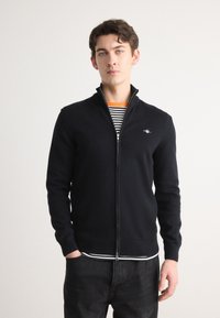 Svart zip-up genser i myk strikket materiale, med høy krage, ribbestrikkede ermer, og en liten brodert logo på brystet.