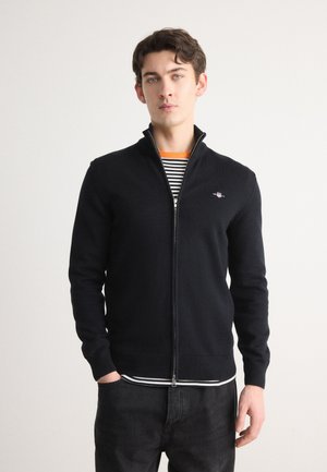 Schwarze Zip-Up-Umhangjacke aus weichem Strickmaterial, mit hohem Kragen, gerippten Bündchen und einem kleinen gestickten Logo auf der Brust.