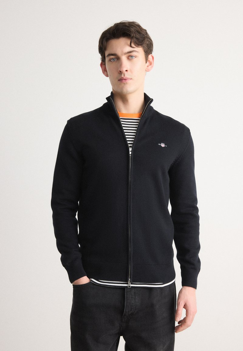 Svart zip-up genser i myk strikket materiale, med høy krage, ribbestrikkede ermer, og en liten brodert logo på brystet.
