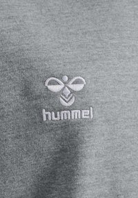 Tessuto di cotone grigio con una consistenza morbida, caratterizzato da un logo ricamato in bianco di Hummel composto da forme astratte e testo sottostante.