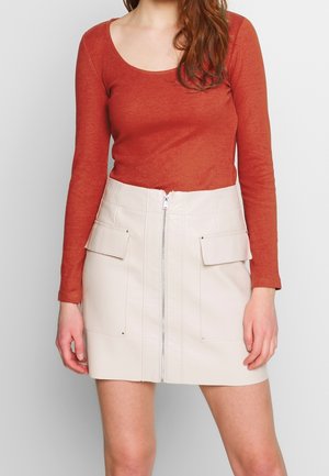 Vrouw draagt een roestkleurige longsleeve en een witte minirok met hoge taille, voorrits en klepzakken.