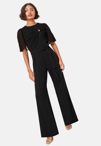 Černý overal s krátkými průsvitnými rukávy, přiléhavým živůtkem se zkrouceným detailem a širokými nohavicemi. Obsahuje páskový vázání a zlaté kovové akcenty.