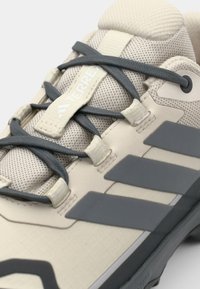 Zapato de senderismo beige con parte superior de malla, cordones grises y tres rayas grises en el lado. Cuenta con una suela texturizada y una lengüeta acolchada para mayor comodidad.