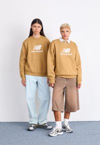 Dva modela nose oversized bež dukseve s logom "New Balance". Jedan ih kombinira s svjetloplavim trapericama, dok drugi nosi smeđe kratke hlače i tenisice.