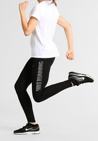 Camiseta deportiva blanca, diseño ajustado, con mangas cortas. Leggings negros con la palabra "RUNNING" en texto reflectante en el lateral. Zapatillas de correr negras.