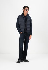 Mann som har på seg solbriller, en marineblå quiltet vest over en marineblå skjorte, mørke jeans og svarte loafers med dusk, står mot en hvit bakgrunn.