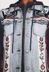 Veste en denim avec broderie florale en rouge, jaune et vert sur le devant, dotée de boutons en argent et d'une capuche grise zippée en dessous.