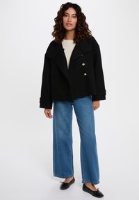 Giacca di lana nera con un ampio colletto e bottoni doppi, abbinata a jeans a gamba larga blu chiaro e ballerine nere.