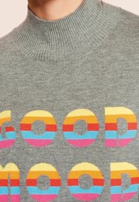 Maglione grigio con colletto alto presenta un testo multicolore che dice "GOOD MOOD" in motivi a strisce giallo, arancione, rosa e blu.