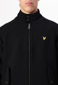 Schwarze Zip-Jacke mit hohem Kragen, ausgestattet mit einem gelben, gestickten Logo. Hergestellt aus strukturiertem Stoff, mit zwei vorderen Taschen und Knopfakzenten.