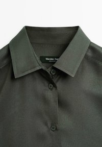 Camicetta verde scuro con colletto, caratterizzata da una texture liscia, chiusura con bottoni e etichetta del marchio all'interno del colletto.