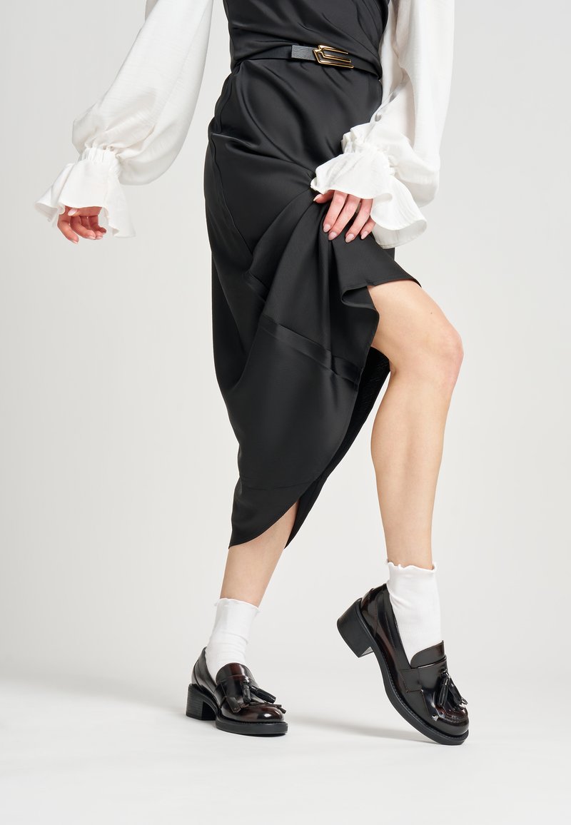 Vestido negro asimétrico con acabado satinado, combinado con mocasines marrón oscuro, calcetas blancas con volantes y un cinturón con hebilla dorada como acento.