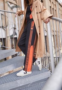 Brauner Trenchcoat über einem schwarzen Trainingsanzug mit orangen und braunen Streifen an den Seiten, kombiniert mit weißen Turnschuhen mit einem dunklen Logo, auf metallischen Treppen.