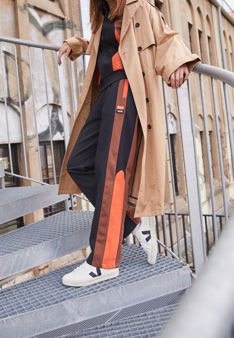 Brauner Trenchcoat über einem schwarzen Trainingsanzug mit orangen und braunen Streifen an den Seiten, kombiniert mit weißen Turnschuhen mit einem dunklen Logo, auf metallischen Treppen.