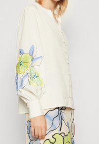Camicia bianca con maniche lunghe a sbuffo, caratterizzata da ricami floreali blu e verdi sulla manica sinistra. Visibili i bottoni frontali e i dettagli dei polsini.